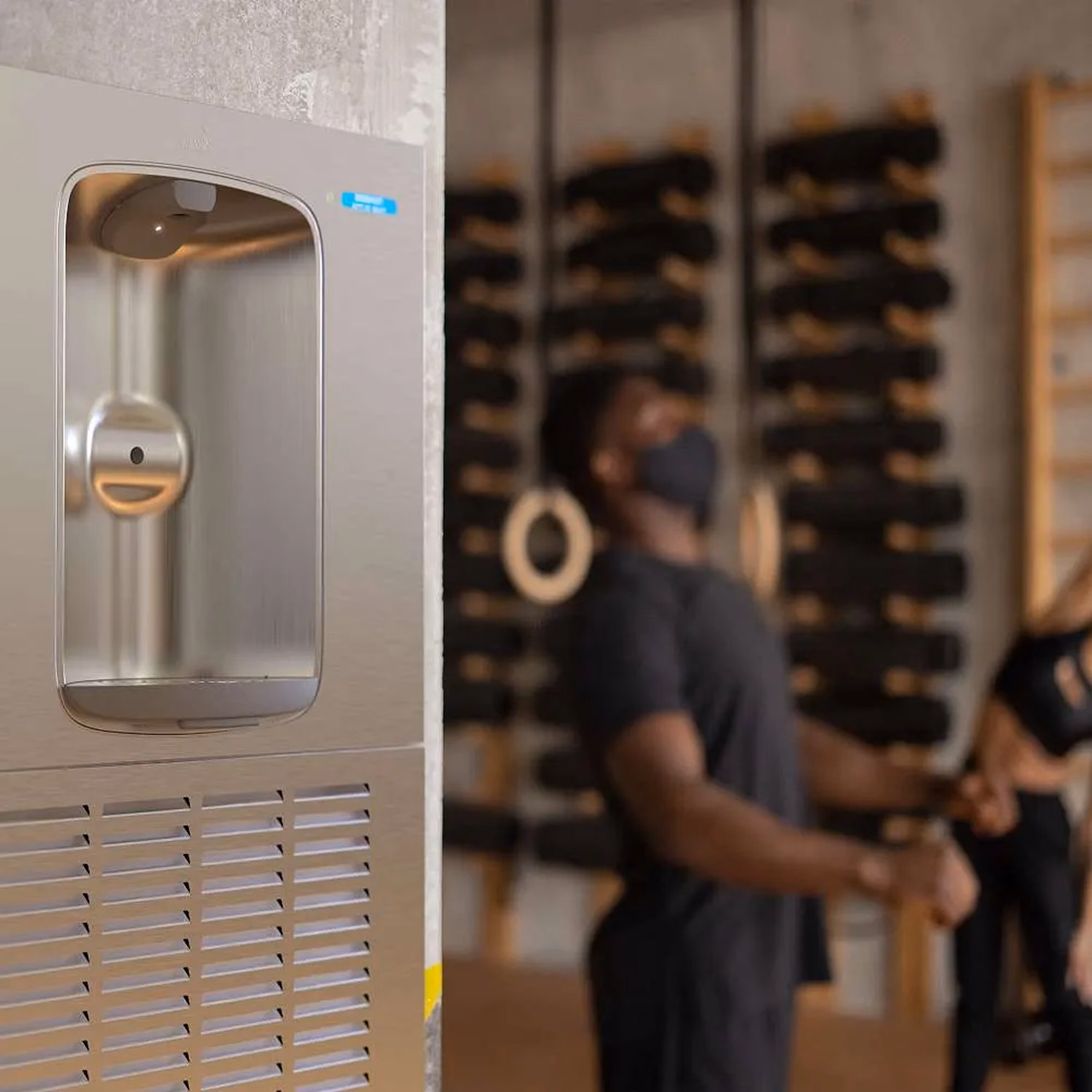 Close-up of a modern stainless steel water dispenser with a blurred background of two people in a gym setting with fitness equipment and gymnastic rings.